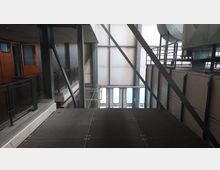 Industrieller Innenraum mit Metallplattform, Gitterboden und sichtbaren Stahlträgern. Im Hintergrund sind ein großer, heller Wandbereich mit Fenstern und Lüftungsanlagen zu sehen, links eine orangefarbene Kabine.