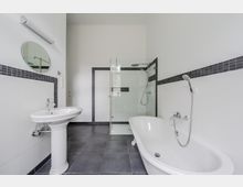 Modernes, helles Badezimmer mit freistehender Badewanne, Waschbecken mit rundem Spiegel und begehbarer Dusche mit Glasabtrennung. Weiße Wände mit dunklem Fliesenband und graue Bodenfliesen prägen den Raum.