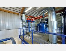 Industrieanlage mit metallischen Rohren, großen Ventilen, einem blauen Druckbehälter und roten Leitungen in einem Gebäude mit Wellblech- und Holzträgerdecke. Der Boden besteht aus einem Gitterrost, umgeben von blauen Metallgeländern.