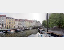 Uferpromenade eines Kanals mit historischen Wohngebäuden im Hintergrund. Mehrere kleine Boote und Schiffe liegen im Wasser, während rechts des Kanals eine Baumreihe und ein Gehweg verlaufen.