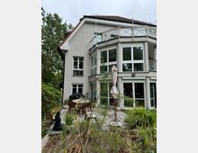 Modernes Wohnhaus mit heller Fassade, großen bodentiefen Fenstern und einem Balkon mit Glasgeländer. Im Vordergrund befindet sich eine Terrasse mit Gartenmöbeln, Pflanzen und einem Sonnenschirm, umgeben von einem angelegten Garten.