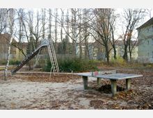 Spielplatz mit einer Metallrutsche und einem Tischtennistisch, umgeben von kahlen Bäumen und Büschen. Der Boden ist mit Sand und herbstlichem Laub bedeckt, im Hintergrund sind Wohngebäude sichtbar.