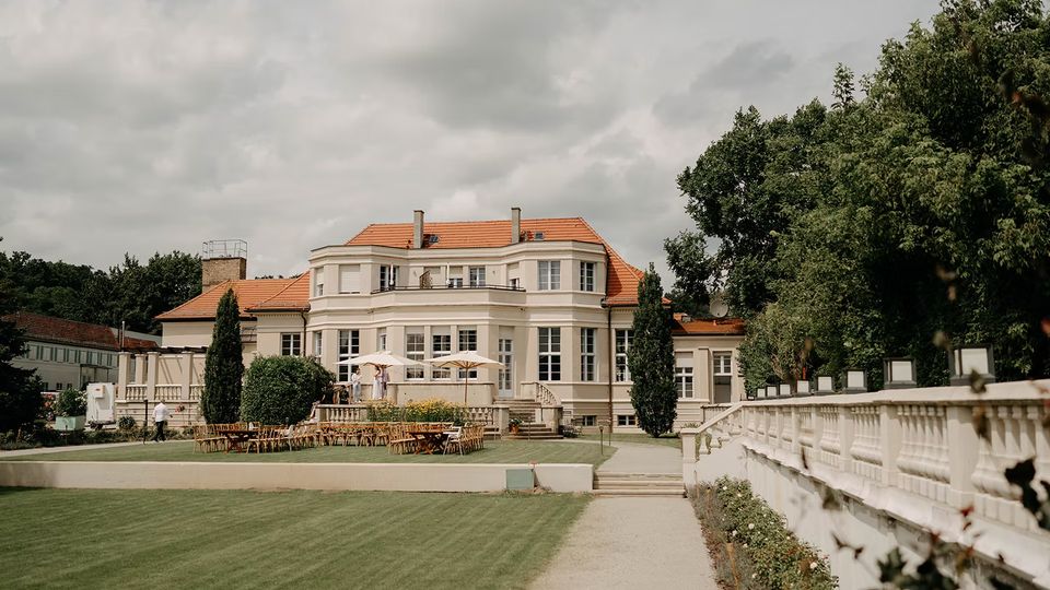 Blick auf eine große, helle Villa mit rotem Ziegeldach und vielen Fenstern, davor eine Terrasse mit Sonnenschirmen und Sitzgelegenheiten. Im Vordergrund führen ein breiter Kiesweg und eine weiße Balustrade durch einen gepflegten Garten mit Rasenflächen und Bäumen.