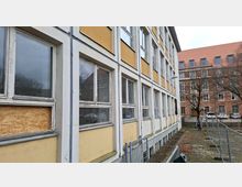 Seitenansicht eines mehrstöckigen Gebäudes mit gelb-weißer Fassade und langen Reihen rechteckiger Fenster, teils beschädigt oder mit Holzplatten abgedeckt. Daneben verläuft ein schmaler gepflasterter Weg mit Metallzäunen; im Hintergrund steht ein größeres Backsteingebäude mit rotem Dach.