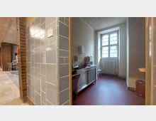 Ein kleiner Innenraum mit einer einfachen Kücheneinrichtung, bestehend aus einer Spüle, einem Wasserhahn, einer Mikrowelle und einem Regal. Der Raum hat rote Bodenfliesen, ein großes Fenster mit Fenstersprossen und einen Heizkörper unter dem Fenster; daneben befindet sich ein Flur mit blauen Fliesen an der Wand.