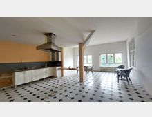 Geräumige Küchen- und Wohnbereich in einem hellen Raum mit schwarz-weißem Fliesenboden, einer weißen Einbauküche mit Metall-Dunstabzugshaube, großen Fenstern und freistehendem Tisch. Im rechten Bereich stehen Rattanmöbel neben einer Fensterfront mit Blick ins Grüne.