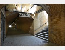 Unterführung mit gelblichen Backsteinwänden, einem gepflasterten Boden und einer Treppe auf der rechten Seite, die nach oben führt. Ein Schild mit der Aufschrift „Gleis 17“ hängt über dem Durchgang.