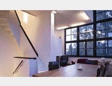 Modernes Wohnraum-Interieur mit hohen Decken, großen Fensterfronten und Blick auf Bäume. Im Vordergrund ein Holztisch mit Stühlen; links führt eine weiße Treppe mit schwarzem Geländer in ein Obergeschoss.