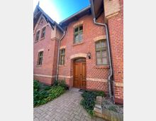 Backsteinfassade eines historischen Gebäudes mit grünen Holzfenstern, einem großen Holzeingangstor und Dachziegeln. Vor dem Eingang befinden sich ein gepflasterter Weg und bepflanzte Beete mit Grünpflanzen.