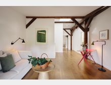 Heller Wohnraum mit weiß gestrichenen Wänden, Holzbalken an der Decke und einem Holzboden. Links steht ein Sofa mit Kissen, daneben ein runder Holztisch mit einer Pflanze in einem Korb, rechts ein kleiner pinkfarbener Tisch mit einer Stehlampe und einem Blumenstrauß.