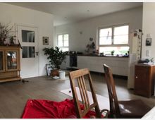 Heller Wohnraum mit offener Küche, weißen Wänden und Holzboden. Im Vordergrund ein Stuhl und ein rotes Tuch, im Hintergrund Küchenschränke, Pflanzen und ein Holzschrank mit Glastüren.