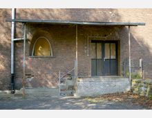 Backsteinfassade eines alten Gebäudes mit verblichenen Doppeltüren, einem zugemauerten Fenster mit Rundbogen, einer kleinen Treppe aus Beton und einem Vordach aus Metall. Daneben sind ein Wasserrohr und ein Metallgitter sichtbar.