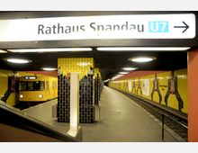 U-Bahn-Station "Rathaus Spandau" mit gelben und schwarzen Wandfliesenmustern, einem gelben U-Bahn-Waggon auf der Linie U7 und einer geschwungenen Bahnsteigkante. Die Decke hat runde Leuchten, die den Bereich beleuchten.