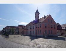 Historisches Rathaus in einem gepflasterten Platz mit hellrosa Fassade, dunkelrotem Ziegeldach und Turmuhr. Umgeben von weiteren kleineren Gebäuden im Fachwerk- und klassischen Stil bei klarem Himmel.