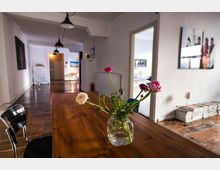 Ein heller, offener Raum mit Holzboden, einem Holztisch im Vordergrund und einem Glasgefäß mit Blumen darauf. Im Hintergrund befinden sich Türen zu weiteren Räumen, Regale mit Zeitschriftenstapeln sowie schlichte Wanddekorationen und Hängelampen.