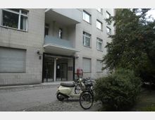 Wohnhausfassade aus hellen Kacheln mit mehreren Fenstern und einem Eingangsbereich mit Vordach. Vor dem Gebäude stehen Fahrräder und ein Motorroller, daneben befinden sich Büsche und ein Baum.