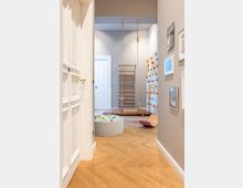 Blick in ein Spielzimmer mit hellem Parkettboden, einer Kletterwand mit orangenen Griffen, einer Sprossenwand mit befestigter Schaukel, sowie einem grauen Bällebad. An den Wänden hängen gerahmte Bilder, im Hintergrund sind weiße Türen zu sehen.