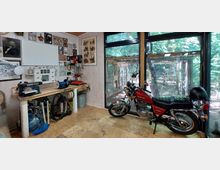 Werkstatt mit einer Werkbank voller Werkzeuge und Maschinen, darunter eine Schleifmaschine und eine Bohrmaschine. Ein rotes Motorrad steht vor einer großen Glasfront, die den Blick auf einen bewaldeten Garten freigibt. An der Wand hängen verschiedene Bilder und Poster.