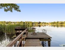 Holzsteg mit Sitzbank und kleinem Tisch führt durch Schilf zu einer Plattform am ruhigen See; seitlich befindet sich eine Metallleiter ins Wasser. Im Hintergrund sind ein bewaldetes Ufer und einzelne Häuser am gegenüberliegenden Seeufer unter klarem Himmel zu sehen.