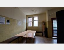 Ein schlichter Raum mit gelblichen Wandfliesen, zwei Fenstern und einer Glastür. Im Vordergrund steht ein Holztisch auf Metallgestell, an der Wand befindet sich eine Bank, und ein zusammenklappbarer Tisch oder Gegenstand lehnt an der Ecke.