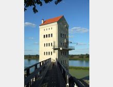 Ein mehrstöckiges Gebäude mit hellem Putz und rotem Satteldach steht am Ufer eines Flusses. Zu dem Gebäude führt ein schmaler Holzsteg, und im Hintergrund sind Wiesen und Bäume sichtbar unter blauem Himmel.