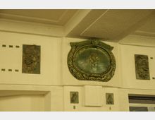 Dekorative Wandgestaltung mit bronzefarbenen Reliefs in einem historischen Gebäude; zentral ein ovaler Rahmen mit Traubenmotiv, flankiert von zwei Fratzenreliefs. Die Wände sind beige gestrichen und mit grünen Akzentdetails verziert.