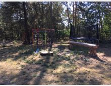 Ein Spielplatz im Freien, umgeben von Bäumen und einem Holzzaun. Zu sehen sind ein buntes Klettergerüst, ein Trampolin, eine Holzbank und ein kleiner Sandkasten auf einem Boden aus Gras und Erde.