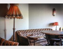 Wohnzimmerartiger Raum mit einer braunen Chesterfield-Ledercouch, einem runden Holztisch, zwei Stühlen und verschiedenen Lampen, darunter eine Stehlampe mit Vintage-Schirm und eine kleine Tischlampe. Die Wände sind schlicht und hell gehalten.