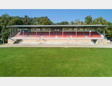 Moderne Tribüne mit rotem Sitzbereich und Überdachung, umgeben von Bäumen. Im Vordergrund befindet sich eine Rasenfläche, während die Tribüne auf einem erhöhten Backsteinsockel mit Treppenaufgängen steht.