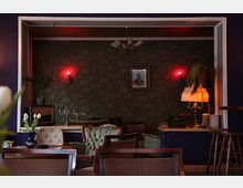 Gemütlicher Innenraum mit Retro-Dekor: Ein Raum mit dunkelgrün gemusterter Tapete, roten Wandlampen, antikem Mobiliar wie Sesseln und einer Kommode. Darauf stehen eine Schreibmaschine, eine Lampe und Pflanzen; ein gerahmtes Schwarz-Weiß-Porträt hängt an der Wand.