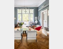 Wohnzimmer mit hellem Parkettboden, einem großen Fenster mit Blick ins Grüne und einer eleganten Einrichtung. Zu sehen sind weiße Sessel, ein roter Stuhl, eine braune Liege mit Fußhocker, ein runder Glastisch und Stuckverzierungen an der Decke.