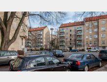 Straßenszene in einem Wohngebiet mit mehrstöckigen, verputzten Gebäuden mit roten Ziegeldächern. Im Vordergrund parken Autos, während im Hintergrund Bäume mit kahlen Ästen und Balkone an den Häusern zu sehen sind.