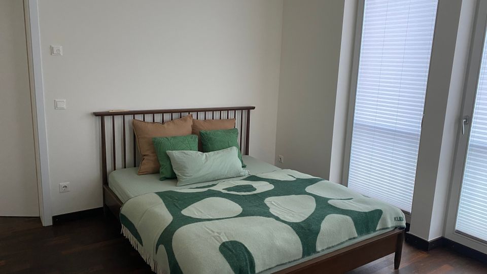 Schlichtes Schlafzimmer mit einem Doppelbett aus dunklem Holz, mehreren Kissen und einer grün gemusterten Decke vor einer hellen Wand. Rechts sorgen bodentiefe Fenster bzw. eine Glastür mit Plissees für Tageslicht; dunkler Holzboden und eine Zimmertür links.