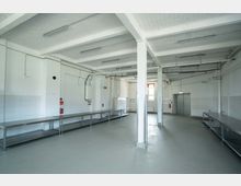 Ein leerer, industrieller Raum mit hellgrauen Böden, weißen Wänden und einer freiliegenden Decke mit Beleuchtung. Entlang der Wände befinden sich mehrere Edelstahltische, sowie Rohre und technische Installationen; im Hintergrund sind ein Fenster und eine doppelte Metalltür erkennbar.