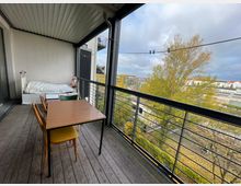 Überdachter Balkon mit Holzboden, einem Tisch und Stühlen, sowie einer weißen Liege auf der linken Seite. Der Balkon bietet Blick auf eine Baumreihe, Eisenbahnschienen und umliegende Gebäude in einer städtischen Umgebung.