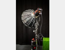 Professionelle Studioleuchte auf einem Stativ mit einem großen, schwarzen Softbox-Aufsatz und Kabelanschlüssen, vor einem schwarzen Hintergrund. Am Boden ist eine Steuereinheit mit roten Akzenten zu sehen.