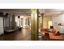 Großer, offener Raum mit Holzfußboden, einer orangefarbenen Ecksofa-Gruppe, freistehenden Metallträgern und großen Fenstern, die viel Licht hereinlassen. Im Hintergrund sind eine kleine Küchentheke, moderne Hängelampen und weitere Sitzmöbel erkennbar.