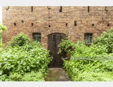 Ziegelsteinfassade eines alten Gebäudes mit einer Holztür in der Mitte, flankiert von zwei kleinen Fenstern mit Sprossenglas. Vor der Fassade wachsen dichte Büsche und grüne Pflanzen.