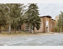 Backsteinbau mit Flachdach und teils vergitterten Fenstern, umgeben von hohen Nadelbäumen; im Vordergrund ein leerer Parkplatz mit markierten Linien.