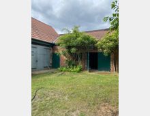 Hof mit einer Ziegeldach-Scheune aus Backstein, grünen Holztoren und umliegenden Bäumen. Der Rasen im Vordergrund ist teilweise bewachsen, während das Wetter bewölkt erscheint.