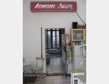 Ein kleines, gemütliches Café im Vintage-Stil mit einem weißen Raum, einer Türöffnung zu einem hinteren Bereich mit großen Fenstern und Holzdielenboden. Im Vordergrund stehen ein runder Tisch, ein Klappstuhl und ein Drehständer mit Postkarten; rechts ist ein Küchenschrank und ein Kühlschrank sichtbar. Ein neonbeleuchtetes Schild mit der Aufschrift „komm rein“ hängt über der Tür.