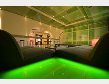 Moderner Lounge-Bereich mit niedrigen schwarzen Sofas um einen Glastisch, darunter eine grün leuchtende Bodenfläche. Im Hintergrund sind eine Bar mit Hockern und eine Wand mit rechteckigen grünen und weißen Mustern zu sehen, ergänzt durch eine abgehängte Decke mit ähnlichem Design.