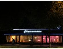 Außenansicht eines Restaurants bei Nacht mit dem Namen „Supersonico“ in leuchtendem Schriftzug. Große Fensterfronten zeigen ein stimmungsvoll beleuchtetes Interieur mit Sitzgelegenheiten, dekorativen Lampen und einem Tresen; davor stehen zwei Personen am Gehweg.
