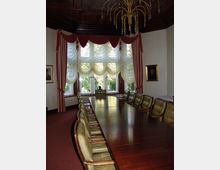 Tagungsraum mit einem langen, polierten Holztisch und gepolsterten Stühlen. Große Fenster mit roten Vorhängen und weißen, gerafften Gardinen lassen Tageslicht herein; an der Decke hängt ein goldener Kronleuchter, und Wände sind mit gemusterten Tapeten dekoriert.