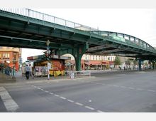 Straßenszene mit einer erhöhten, grünen Stahlbrücke, unter der sich ein kleiner Kiosk befindet. Im Hintergrund sind mehrstöckige Gebäude mit Geschäften, Restaurants und Menschen auf Gehwegen zu sehen.