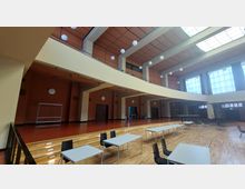 Großer Saal mit modernem Design, hohen Decken und einer Galerieebene. Der Raum verfügt über helle Fensterflächen, eine hölzerne Bühne, Holzfußboden und Tisch-Stuhl-Arrangements, die auf eine multifunktionale Nutzung hinweisen.