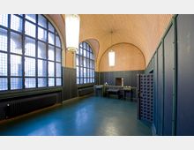 Innenraum eines historischen Gebäudes mit gewölbter Decke, die mit einem wabenartigen Muster verziert ist. Große, bogenförmige Fenster durchfluten den Raum mit Licht; dunkle Holzwände und funktionale Sitzgelegenheiten prägen das Mobiliar.