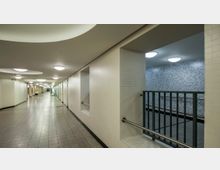 Ein moderner U-Bahn-Korridor mit hellen Fliesenwänden und grauen Bodenfliesen. Rechts befindet sich ein Bereich mit Geländer, der in eine niedrigere Ebene oder Treppe führt, beleuchtet durch gleichmäßig verteilte Deckenleuchten.