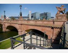 Sandsteinbrücke mit kunstvollen Verzierungen, darunter Greif-Statuen und Laternen, über einem Fluss; im Hintergrund das moderne Glasgebäude des Berliner Hauptbahnhofs. Im Vordergrund ein schmiedeeisernes Geländer einer Treppe.