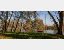 Ufer eines ruhigen Flusses mit Bäumen im Vordergrund und herbstlich gefärbten Bäumen am gegenüberliegenden Ufer. Eine rote Bank steht auf einer grünen Wiese, und ein Strommast ist in der Ferne sichtbar.